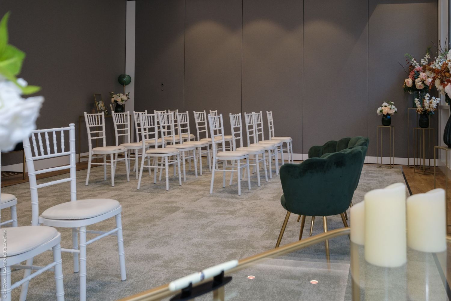 Wedding venue setup with white Chiavari chairs, green velvet armchair, candles, and floral decorations on gray walls.
