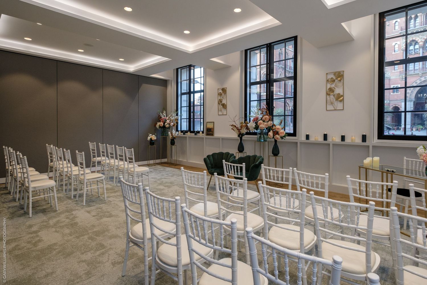 Elegant wedding ceremony room with white Chiavari chairs, large black-framed windows, and floral arrangements on a polished floor.