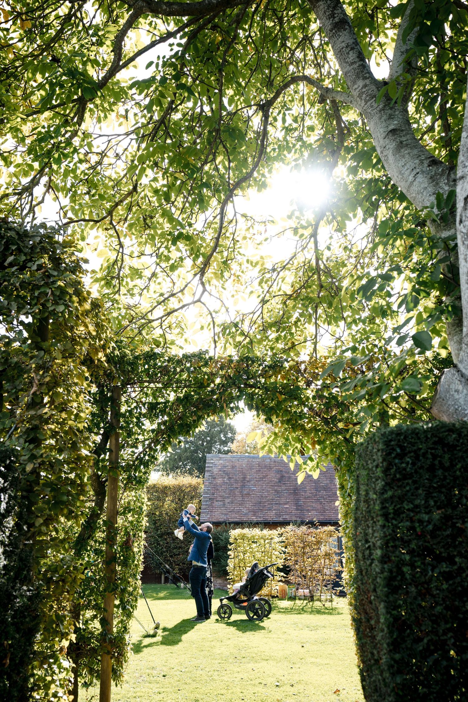 Golfer with trolley on lush green course, framed by hedgerow arch and sunlit tree canopy near rustic building.