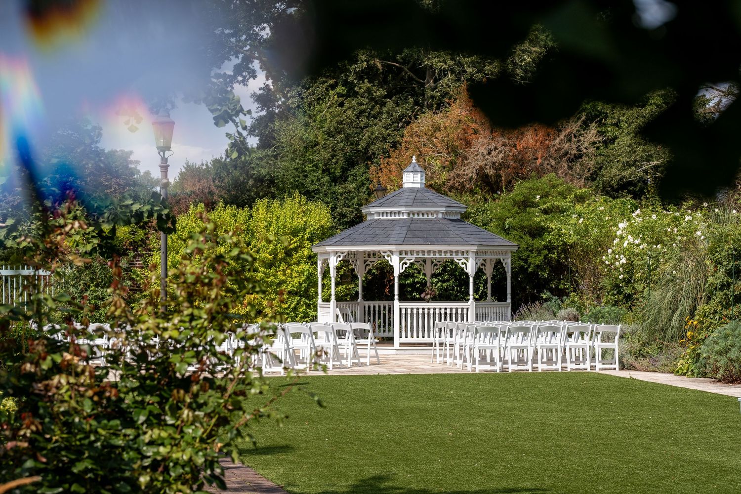White gazebo wedding ceremony setup with chairs on green lawn surrounded by lush gardens and trees on a sunny day.