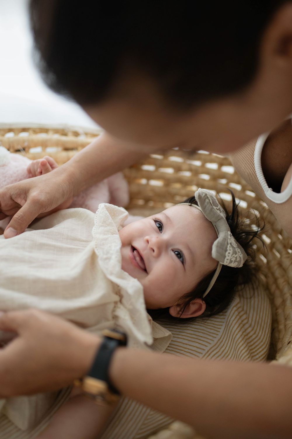 A smiling baby girl in a white ruffle dress and floral headband lying in a wicker basket with an adult leaning over her.