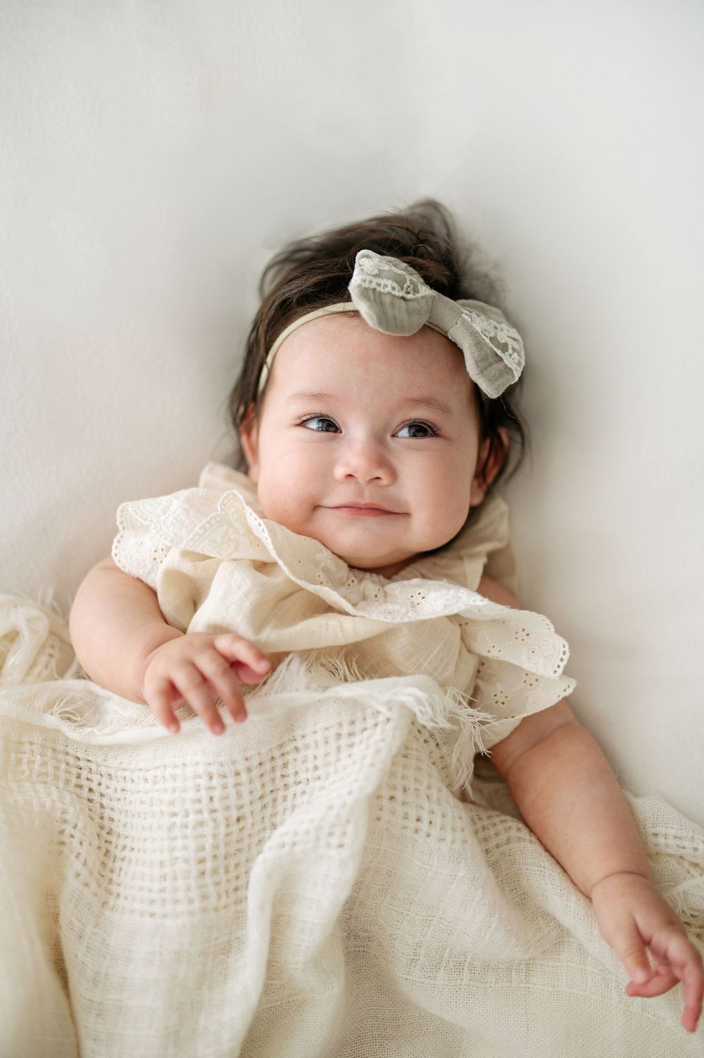 Smiling baby girl in cream eyelet dress and sage bow headband lying on white blanket.