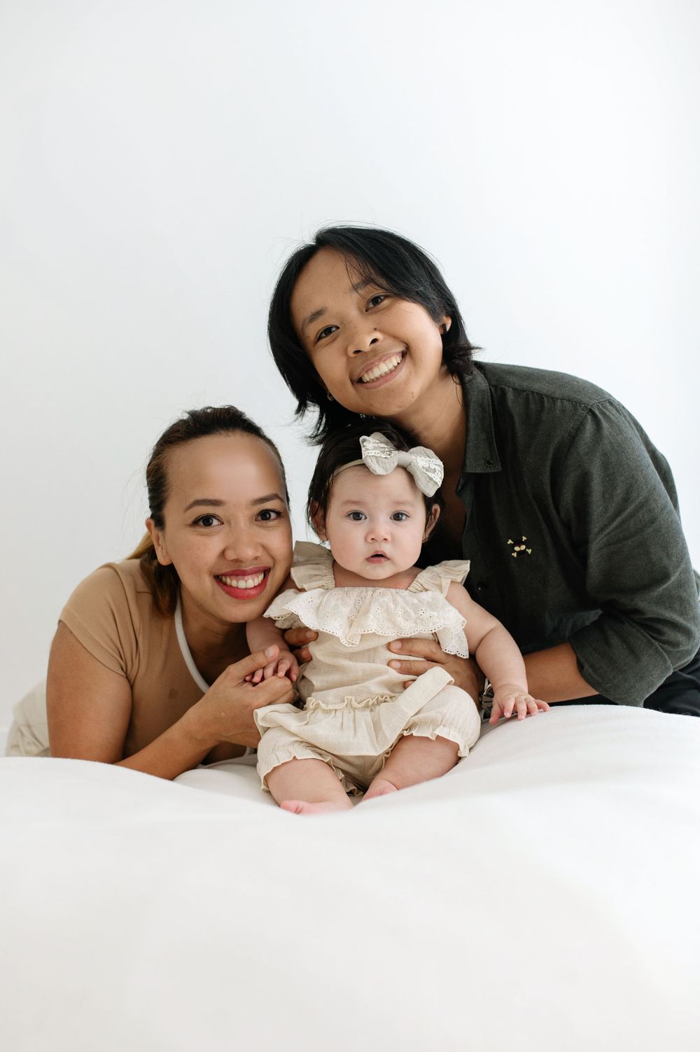 Two smiling women pose with a baby girl in a white bow and cream outfit against a white background.