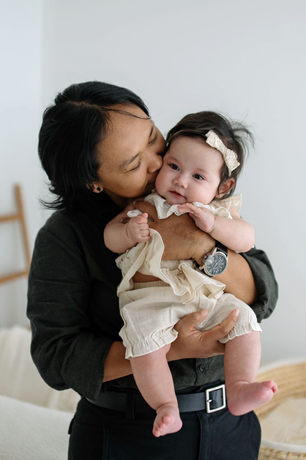 A parent tenderly kisses their baby girl wearing a cream ruffle outfit and bow headband in a bright, minimalist room.
