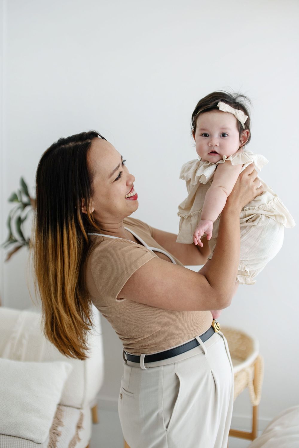 A smiling woman holds up a baby girl in a white dress with a bow headband in a bright, modern room.