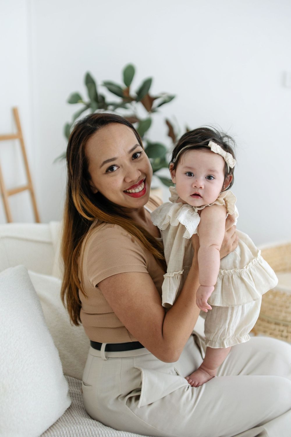 Smiling woman in beige outfit holding an adorable baby girl in a ruffled dress with a bow headband on a white sofa.