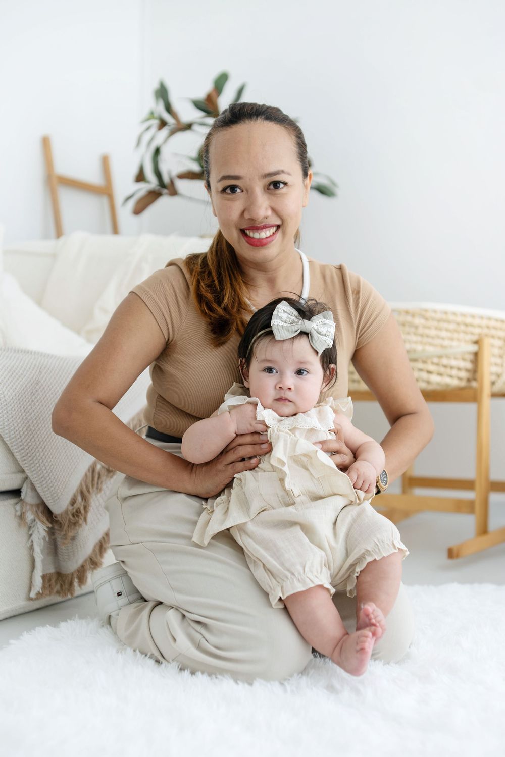 Smiling woman in beige outfit sits on white rug holding an adorable baby girl in cream romper with bow headband.