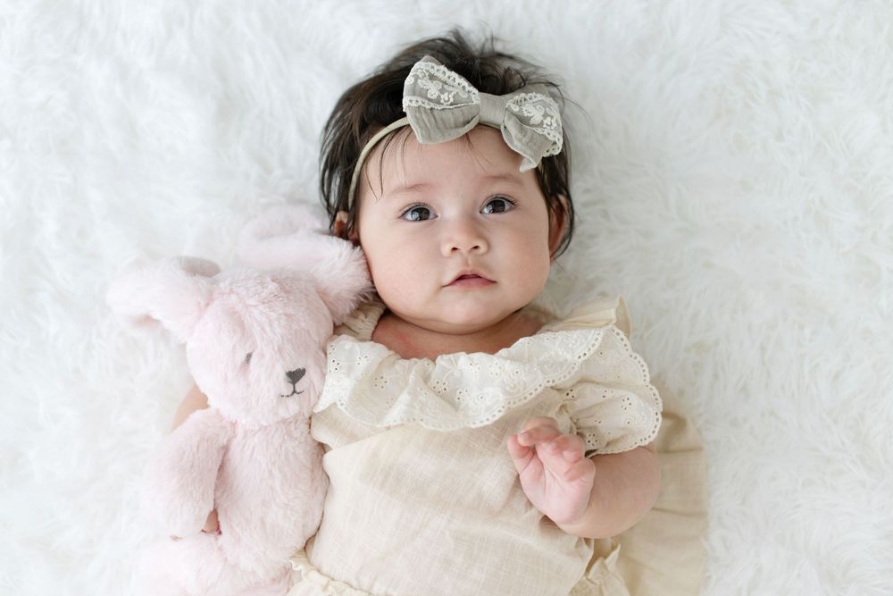 Baby girl in white lace dress with silver bow headband lying on fur blanket holding pink teddy bear.