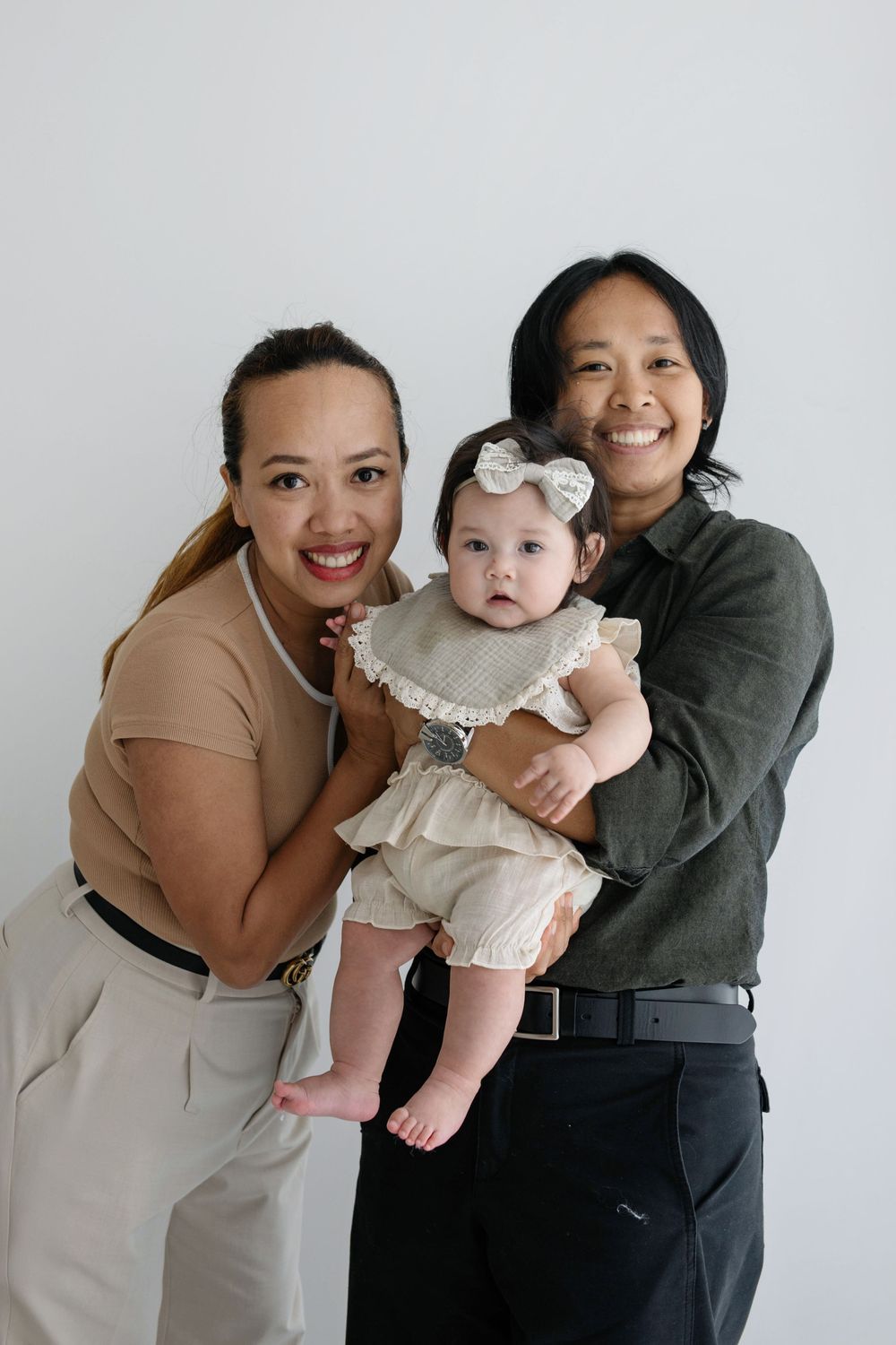 Two smiling parents hold their baby girl in cream romper with bow headband against a white background.