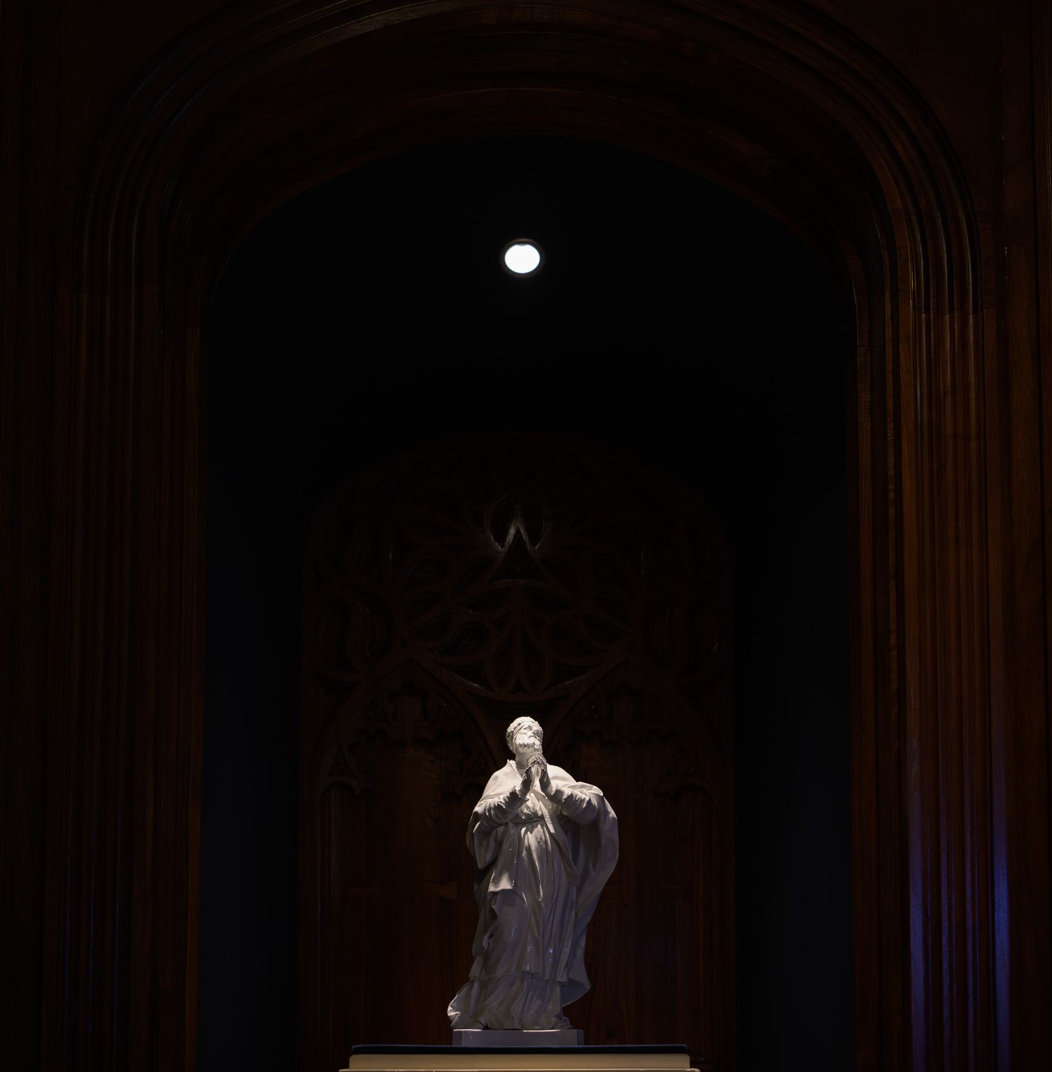 White marble statue of a praying figure illuminated under a glowing orb in a dark arched cathedral alcove.