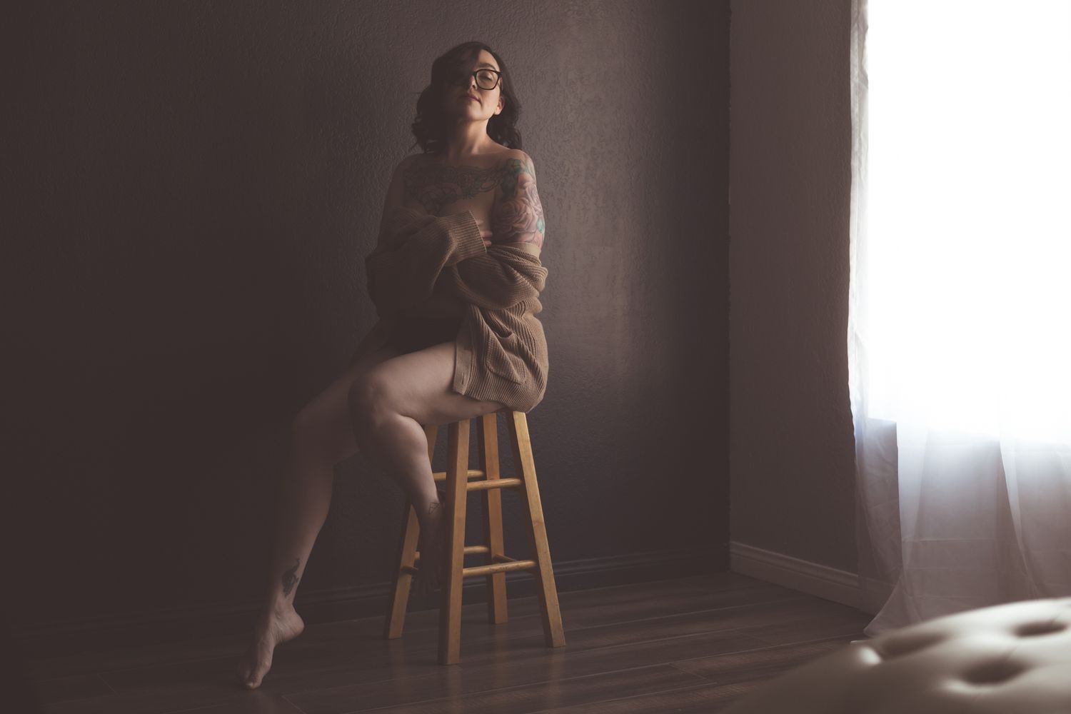 Tattooed woman in white bodysuit sitting on cream sofa, wearing glasses, posing against gray wall.