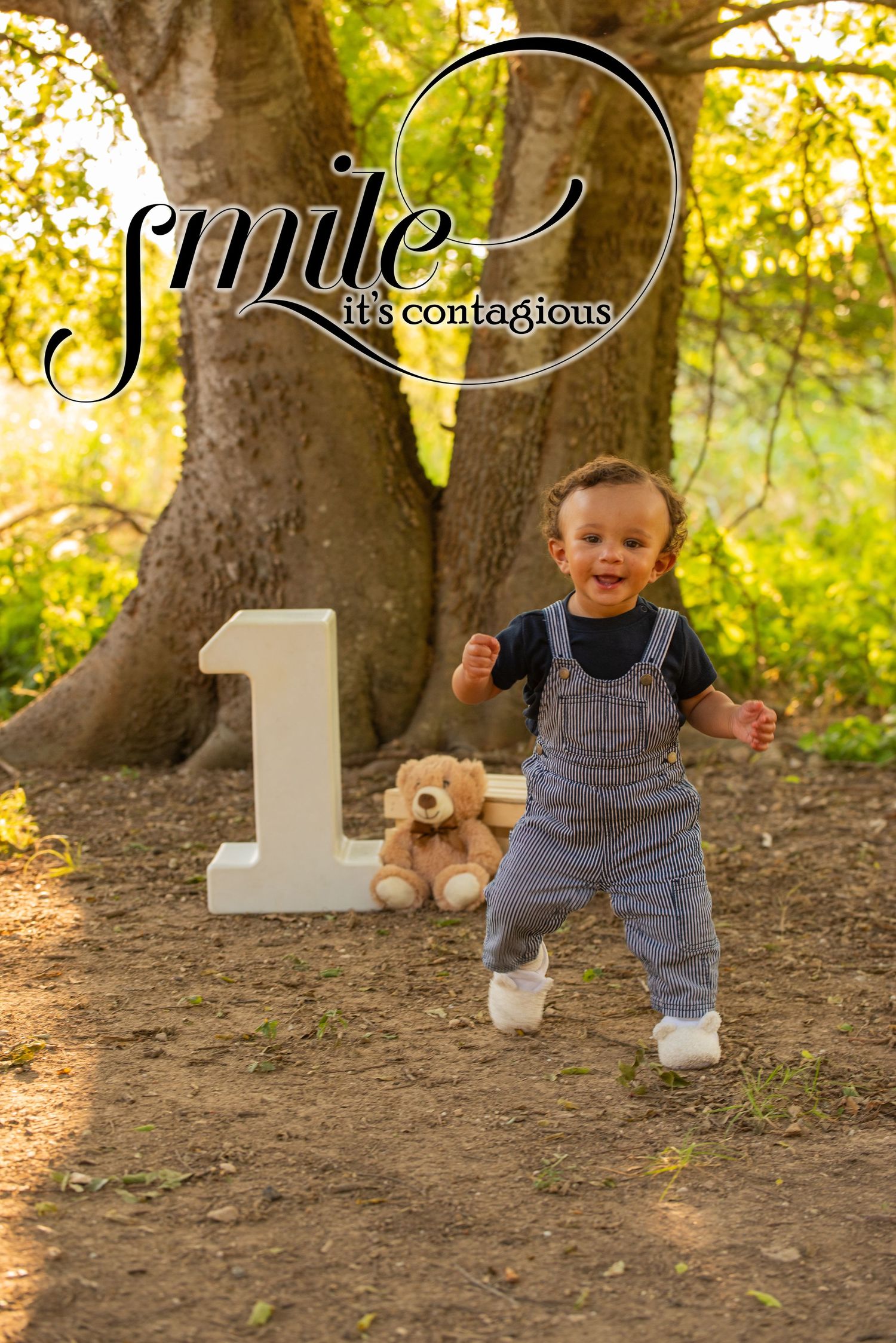 A joyful toddler in denim overalls celebrates his first birthday outdoors near a large tree with a number one prop and teddy bear.