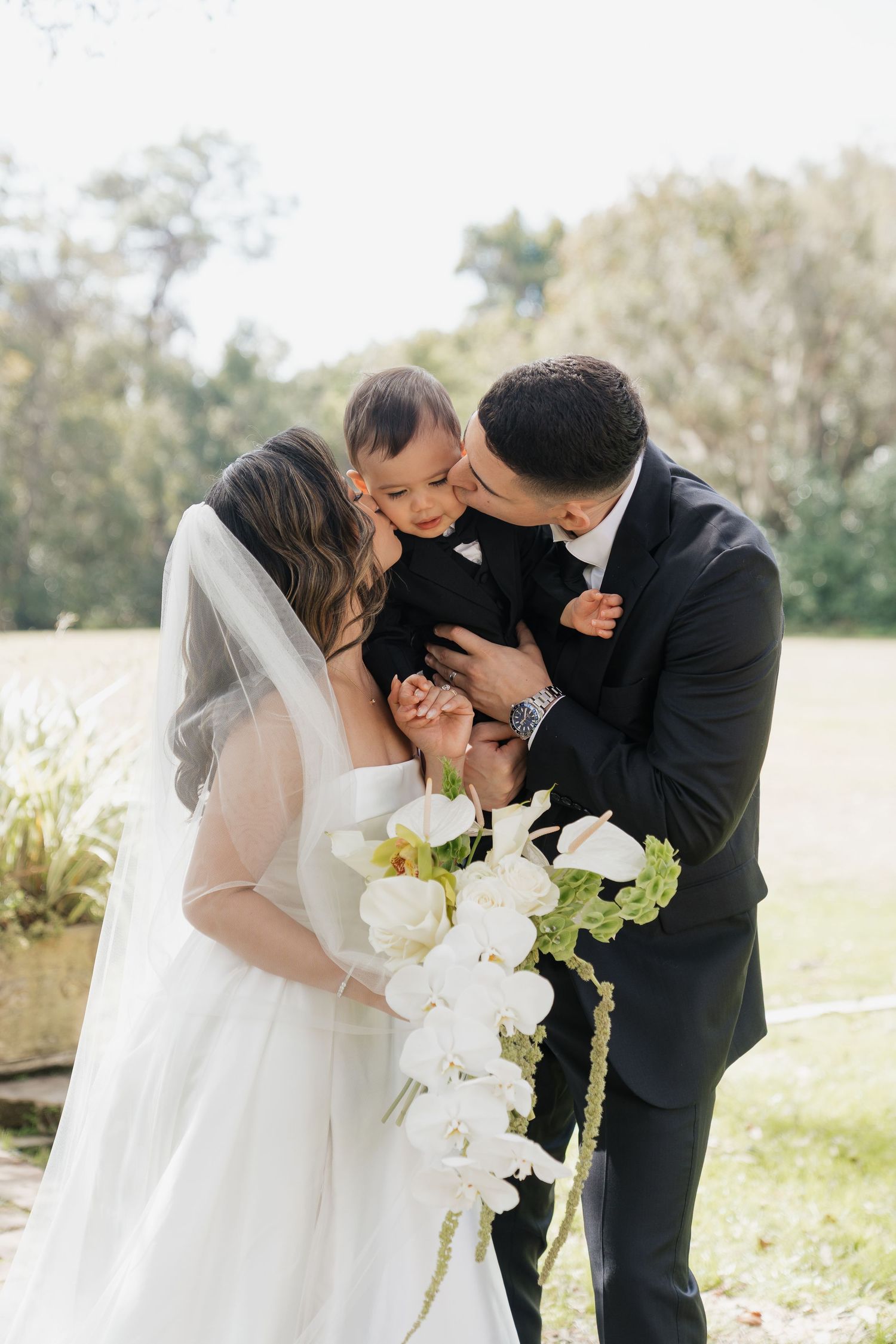 Bride and groom kissing their baby on wedding day, bride holding white orchid bouquet outdoors.