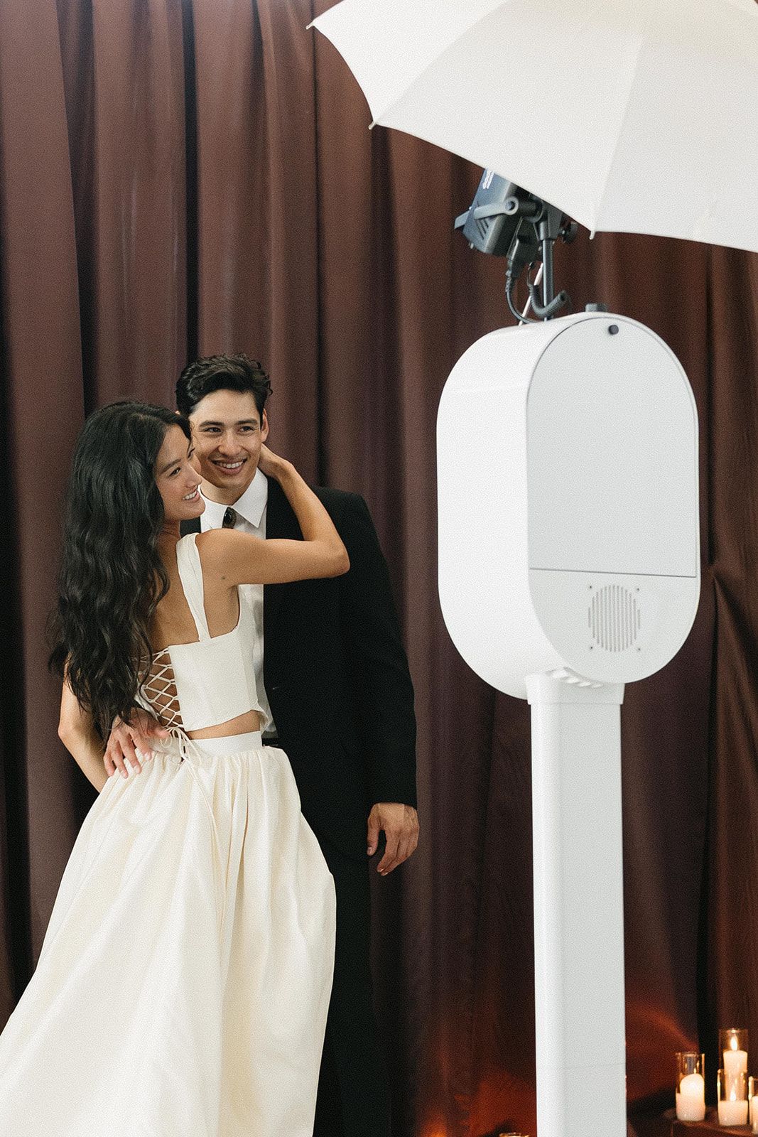 A bride and groom smile together near a photo booth with an umbrella flash at their elegant wedding reception.