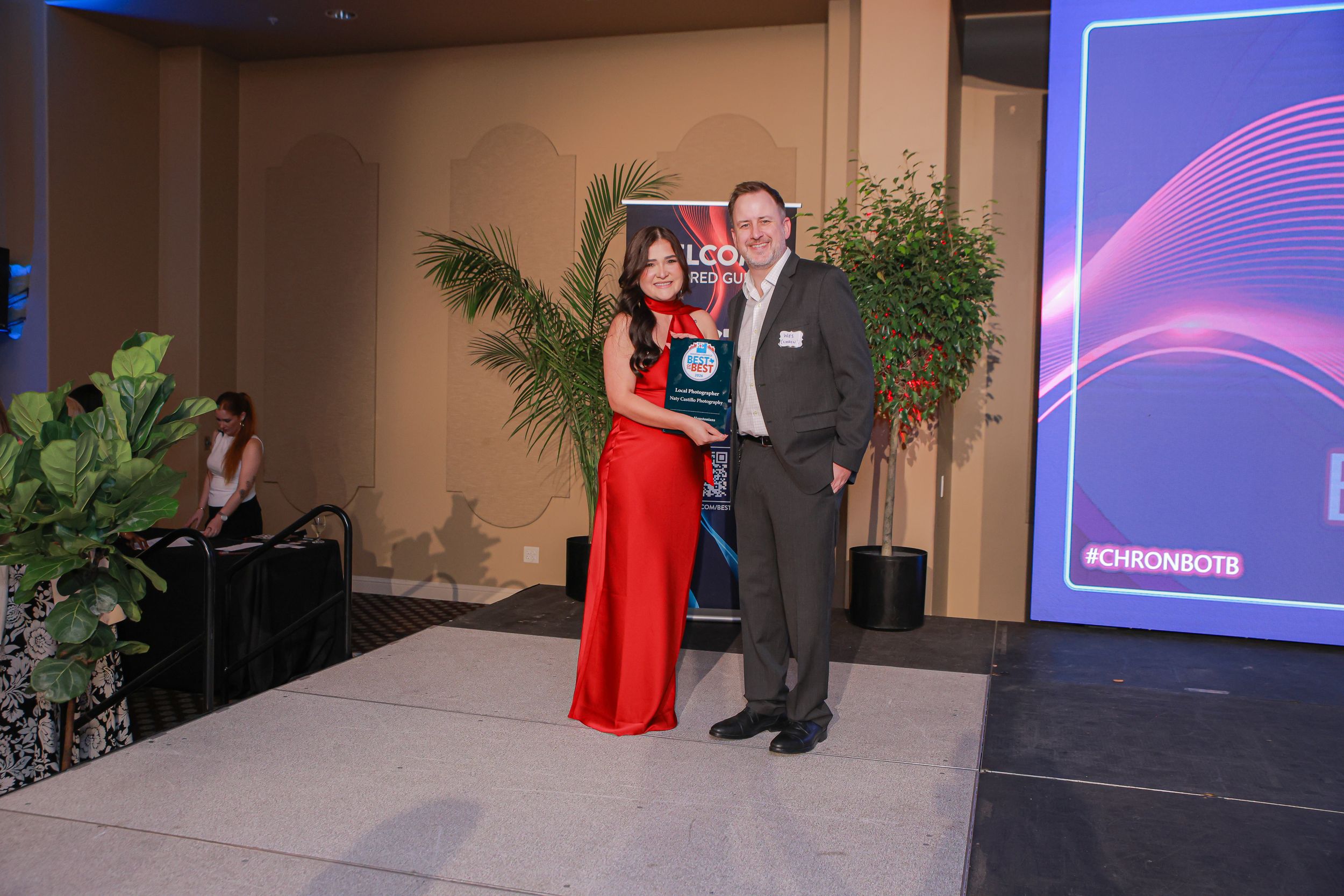 A woman in a red dress and man in a suit pose together on stage at a formal awards ceremony event.