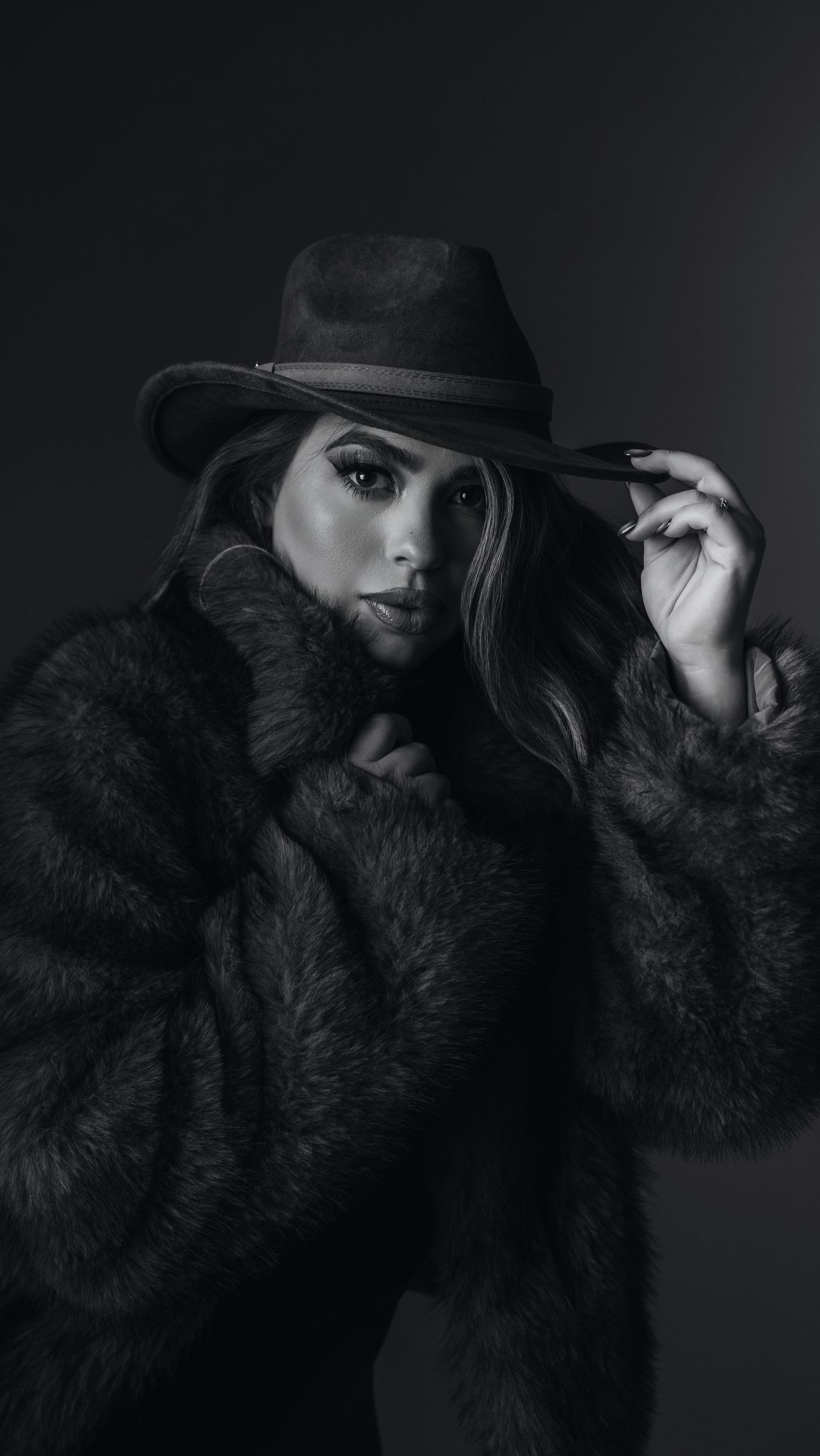 Black and white portrait of a woman in a fur coat and fedora hat, posing elegantly against a dark background.