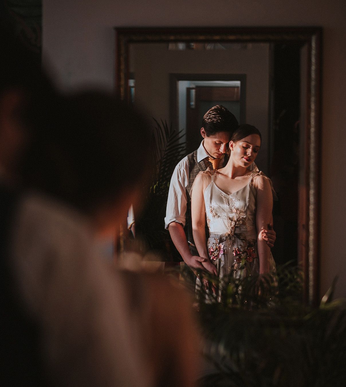 Couple in wedding attire reflected in ornate mirror, surrounded by lush greenery in warm, moody lighting.