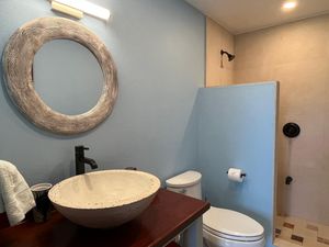 Modern bathroom with round driftwood mirror, vessel sink on wood vanity, blue walls, and tiled shower area.