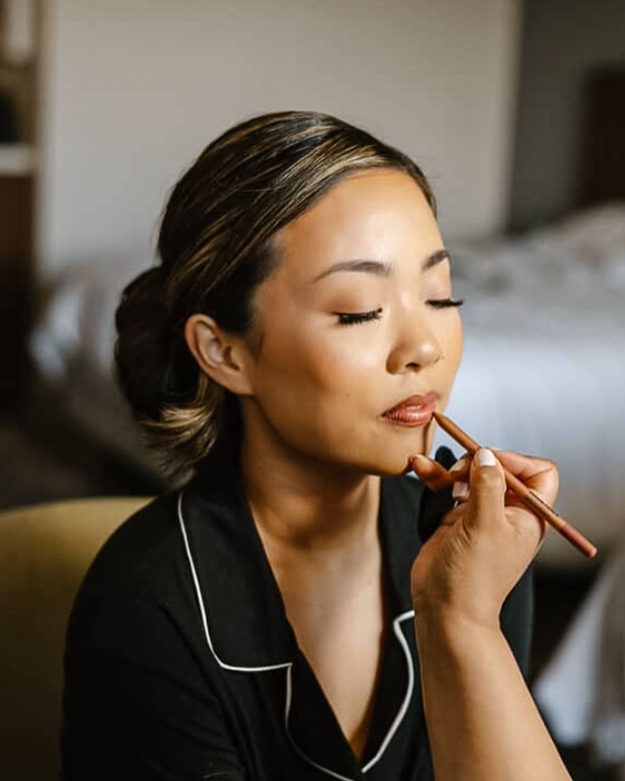 A woman in black satin pajamas gets her lips lined with a nude lip pencil during bridal makeup preparation.