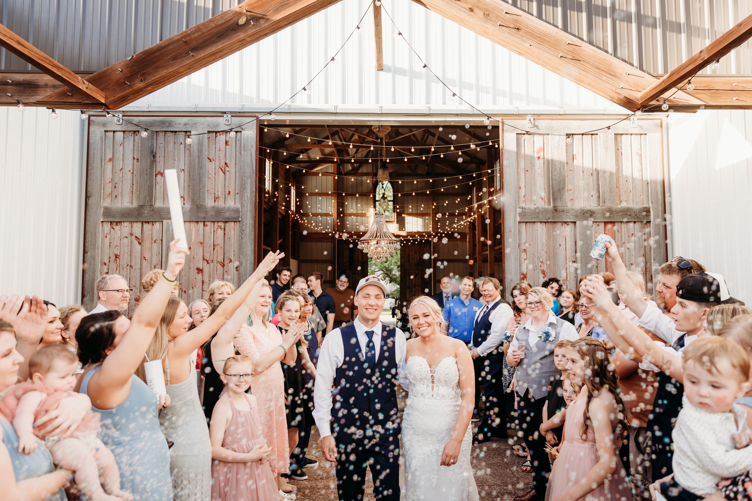 Bride and groom exit barn wedding venue through confetti shower surrounded by cheering guests.