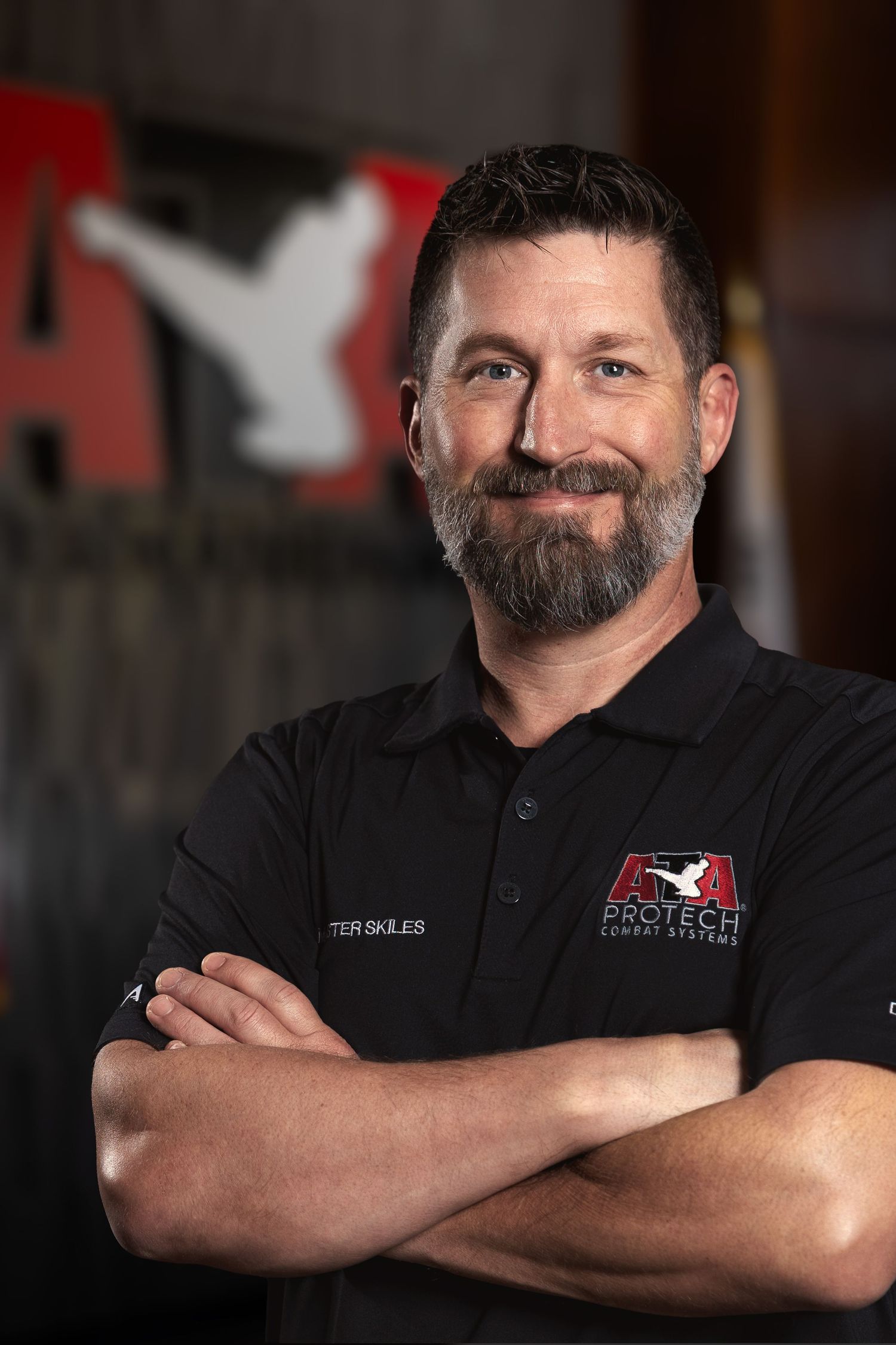 Man in black Protect Combat Systems polo shirt stands with arms crossed, smiling confidently in a martial arts studio.
