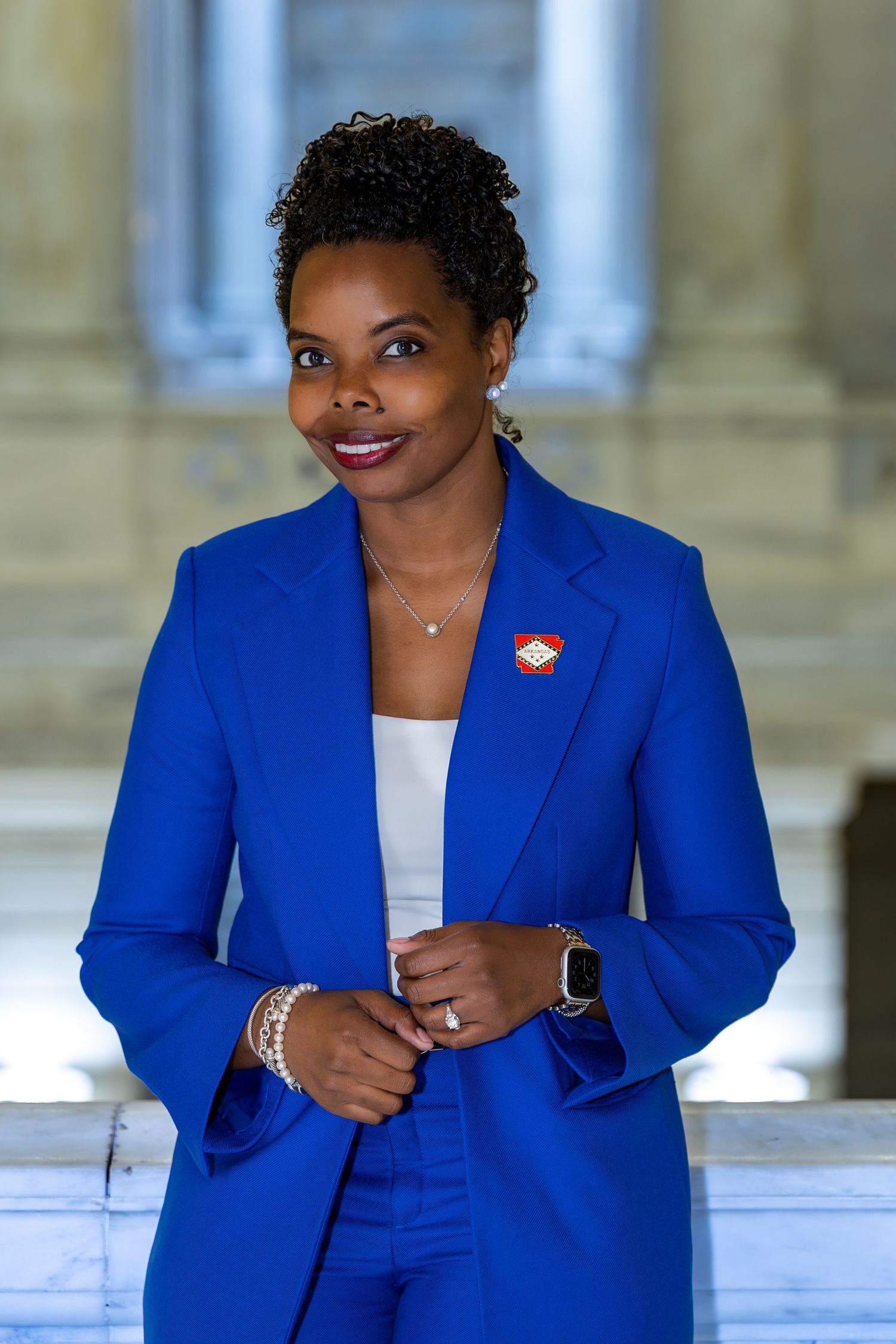 Professional woman in blue blazer and pants smiling indoors, wearing an Arkansas state pin.