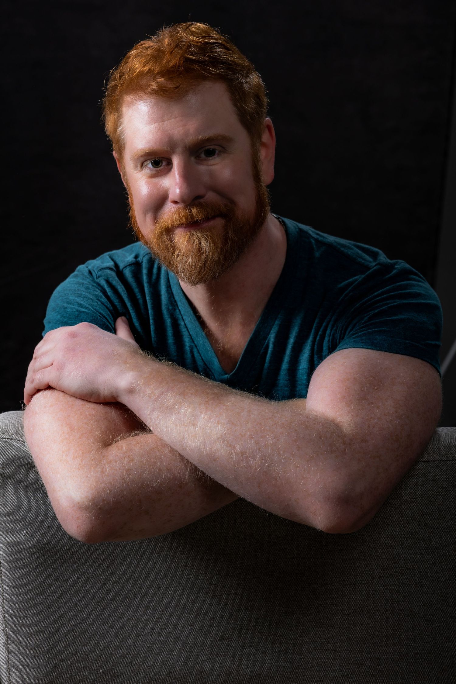 Red-haired bearded man in teal shirt rests arms on sofa back against dark background.