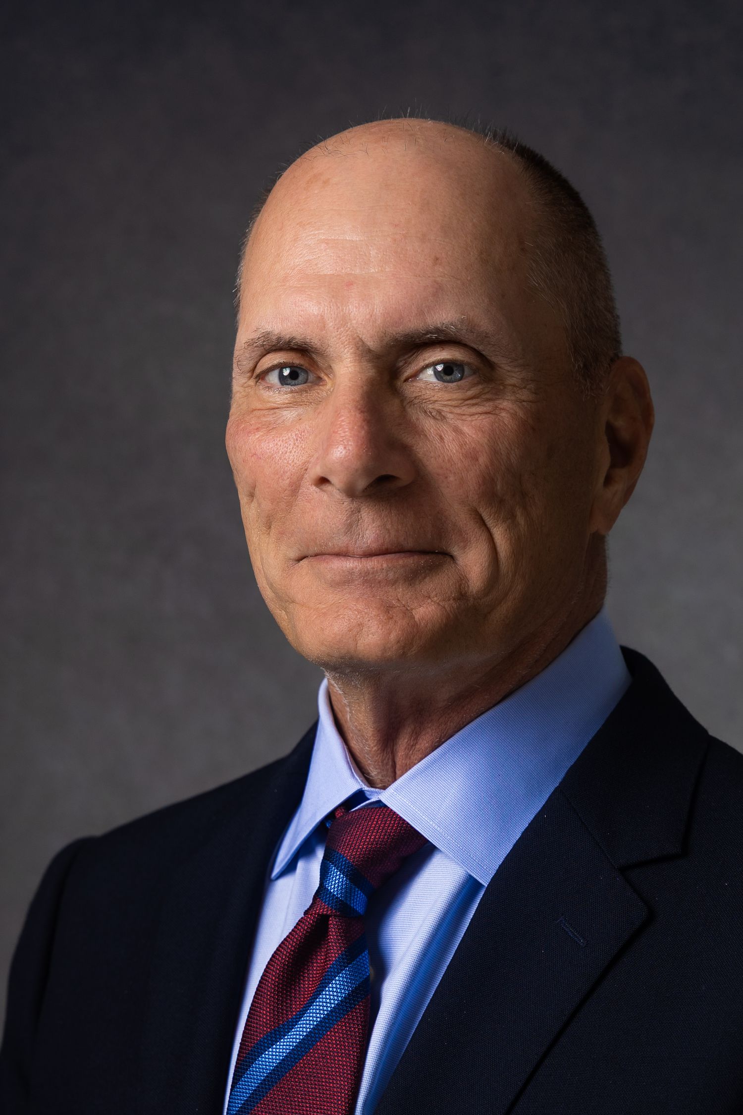 Professional headshot of a bald man in a navy suit and striped tie against a gray background.
