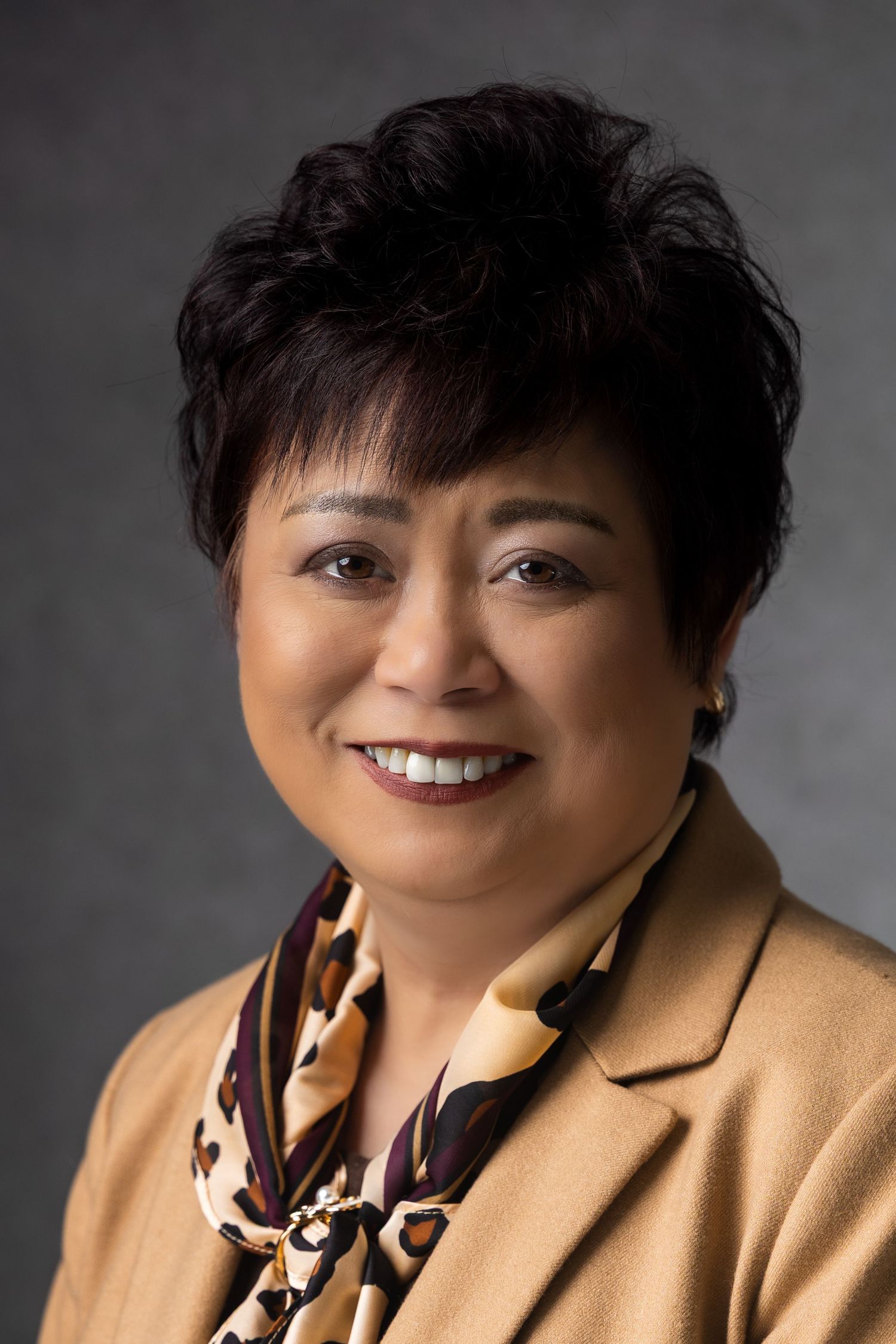 Professional headshot of a smiling middle-aged Asian woman with short dark hair, wearing a tan blazer and floral scarf.