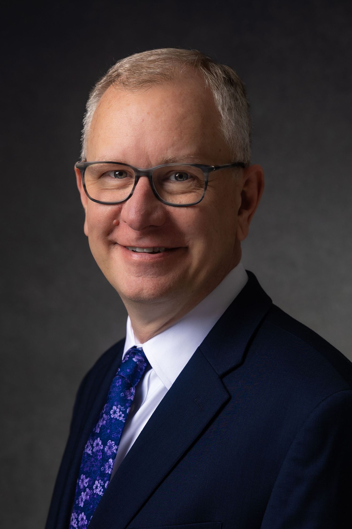 Professional headshot of a smiling middle-aged man with gray hair, glasses, and a navy suit with floral tie.