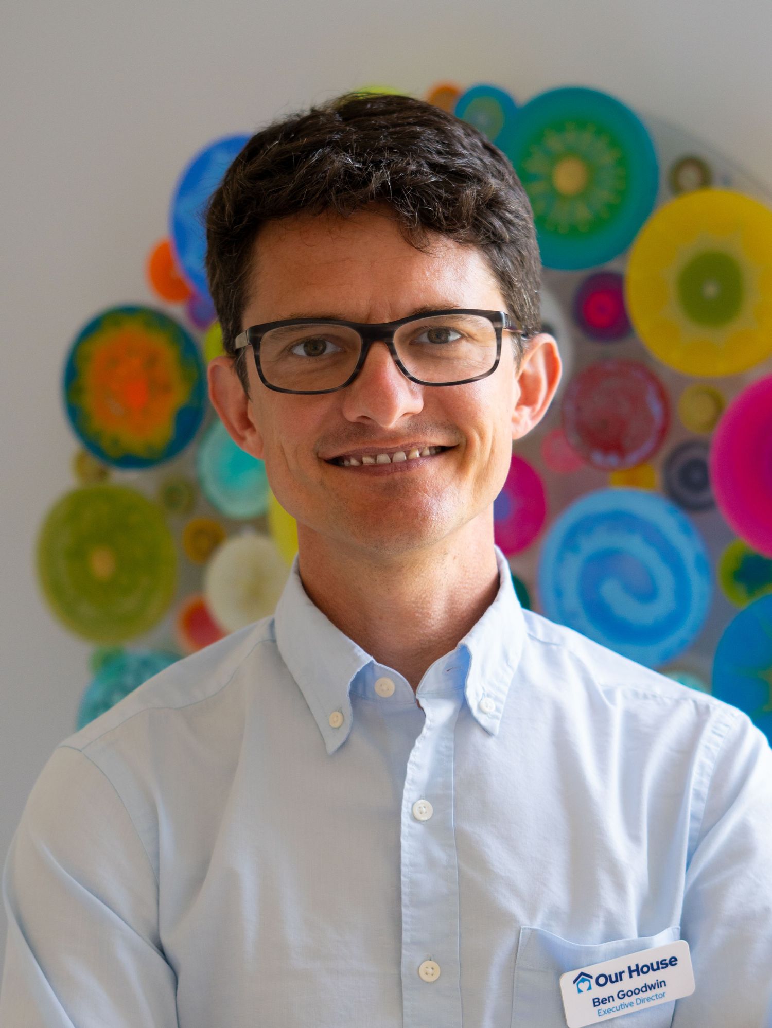 Professional headshot of a smiling man wearing glasses and a blue shirt with an Our House name badge.