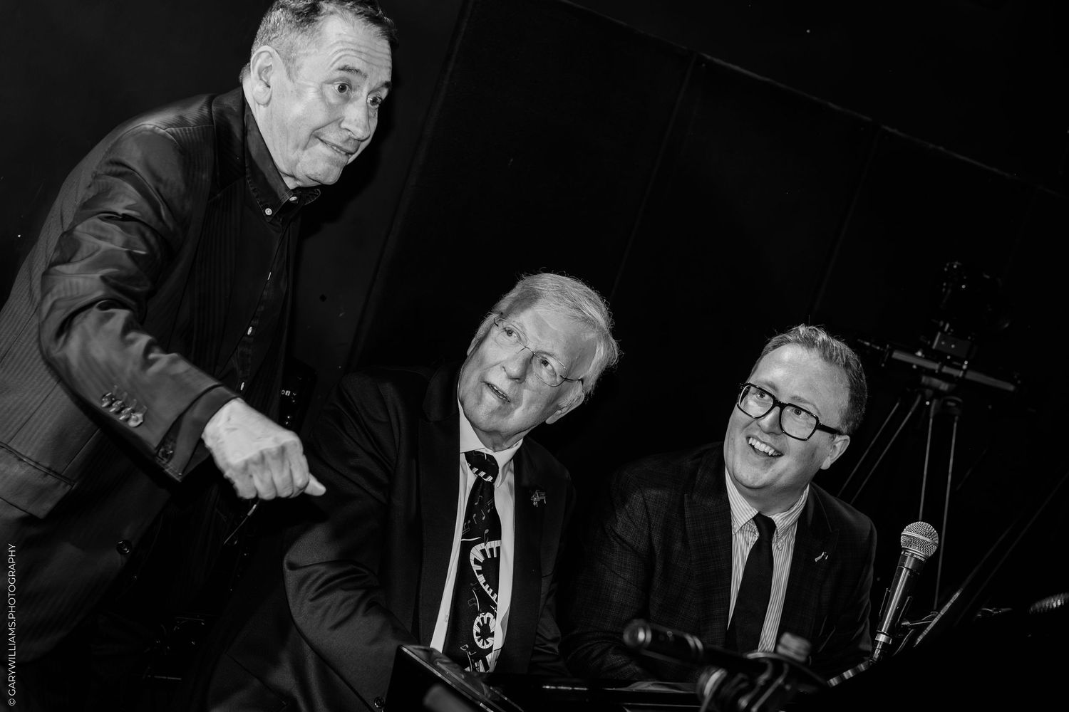Three men in suits smiling and laughing together at a piano in a black and white photo.