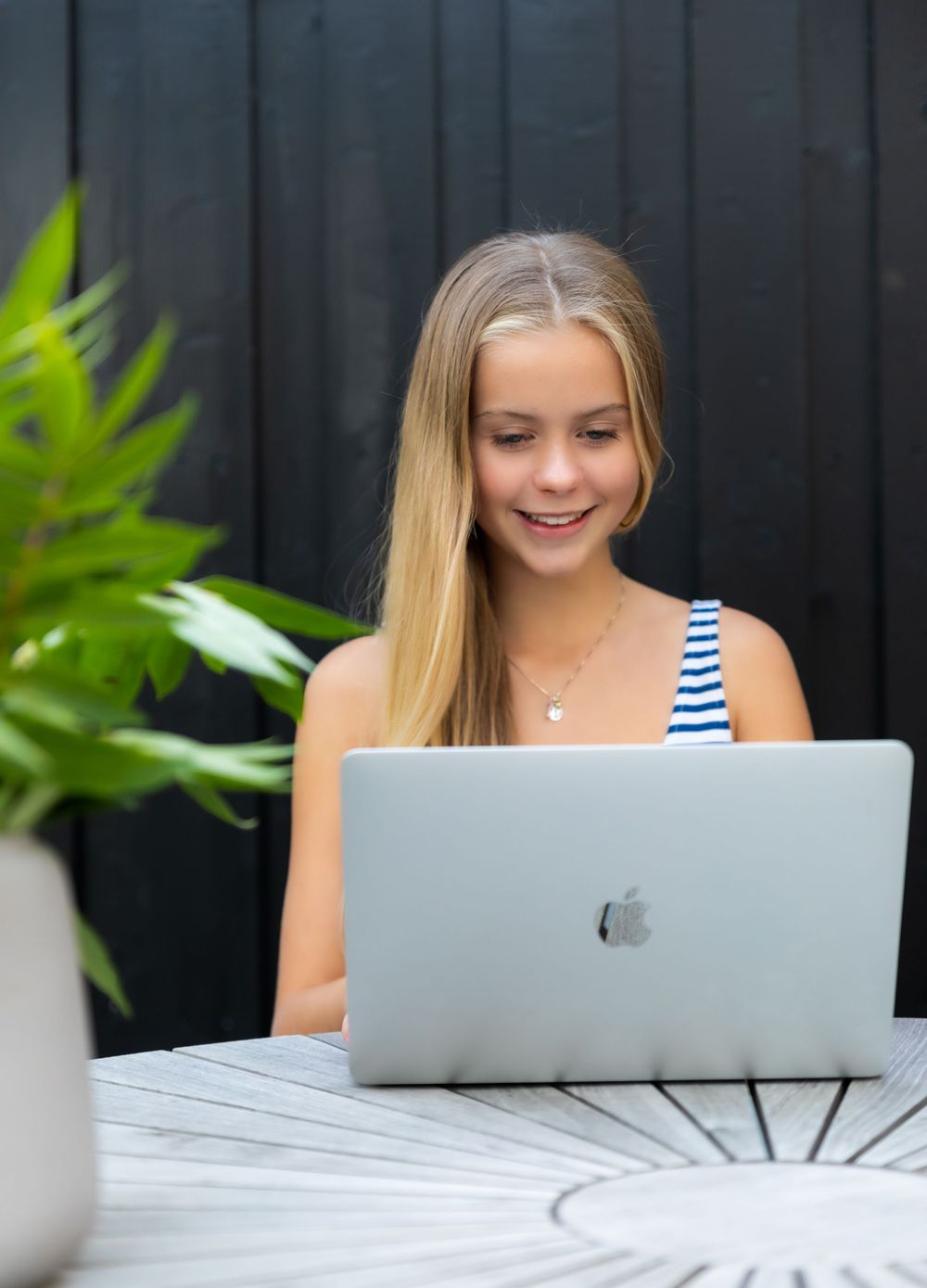 Young blonde girl smiling while using an Apple MacBook laptop at an outdoor wooden table.