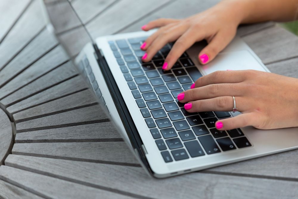 Woman with pink nails typing on a silver laptop at an outdoor wooden table.
