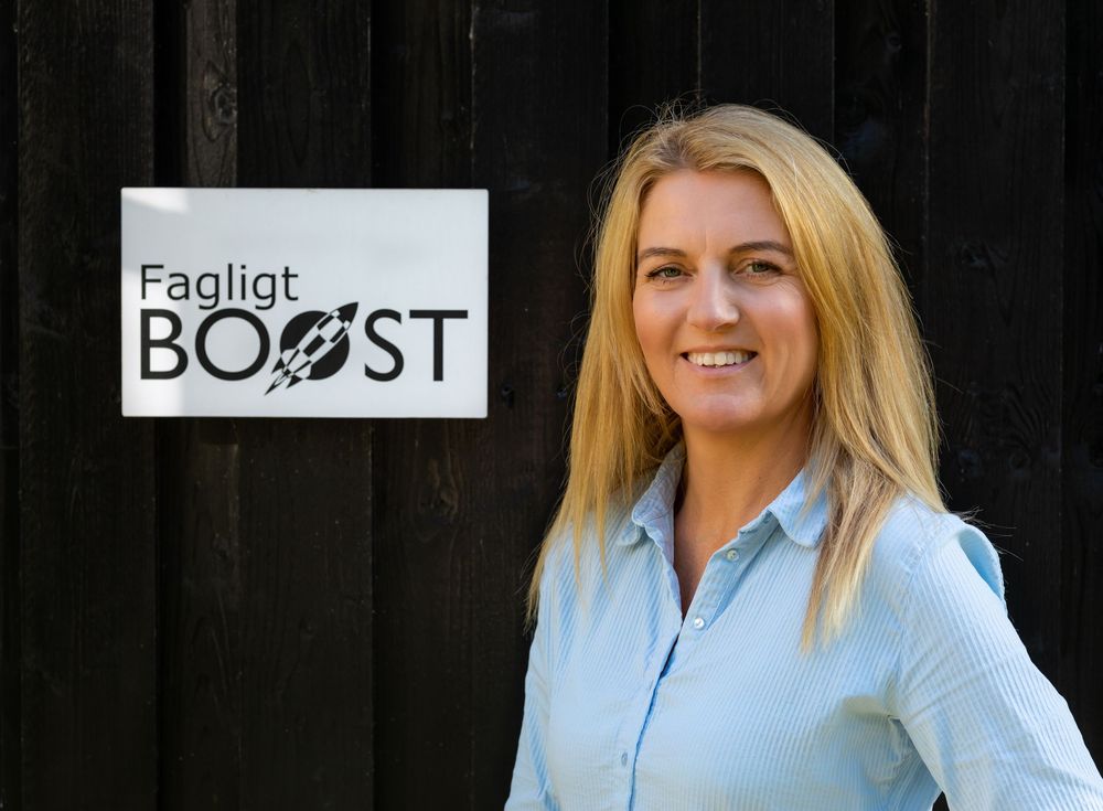 Smiling blonde woman standing next to a Fagligt BOOST sign on a dark wooden wall.