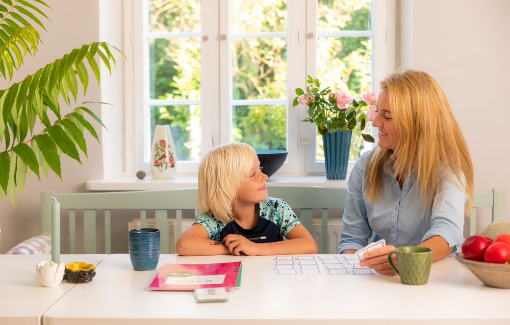 A mother and child sit together at a white kitchen table, smiling and talking in a bright, cozy home.
