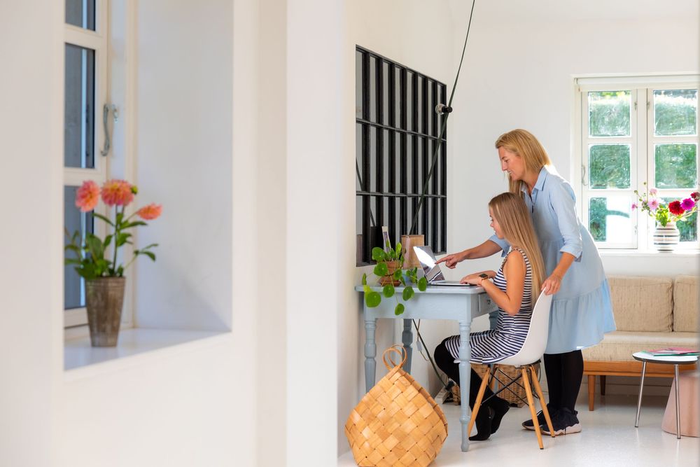 Two women collaborating at a white desk in a bright Scandinavian-style living room with flowers and natural light.
