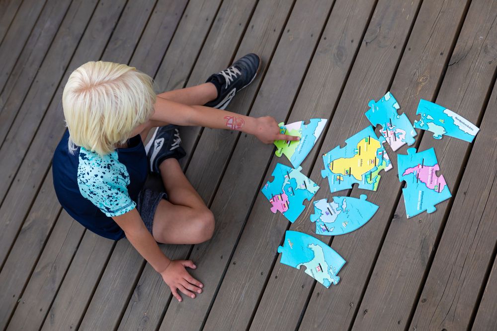 Blonde child sitting on wooden deck playing with colorful puzzle pieces outdoors.