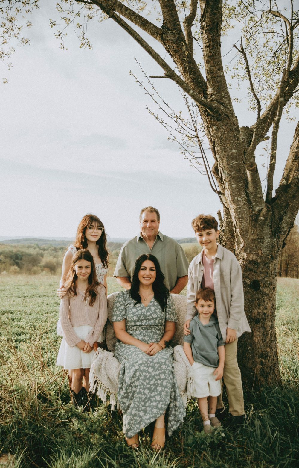 Family of six posing outdoors near a large tree in a green field with rolling hills in the background on a sunny day.