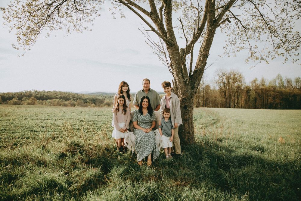 Family of six posing together under a large tree in a lush green field during golden hour.