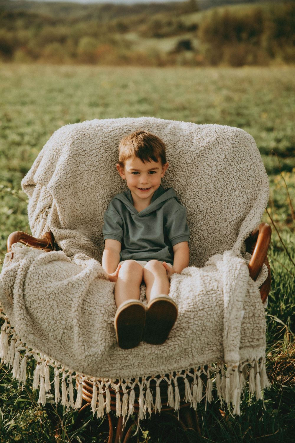 A smiling young boy sitting in a cozy armchair draped with a fringe blanket, outdoors in a green field.