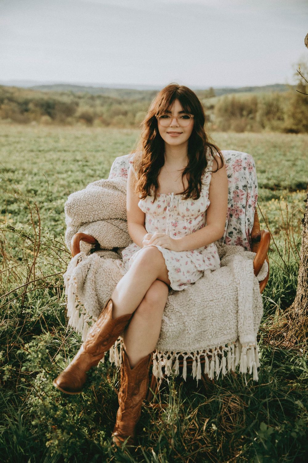 Young woman in floral dress and cowboy boots sitting in armchair in a green field with rolling hills backdrop.