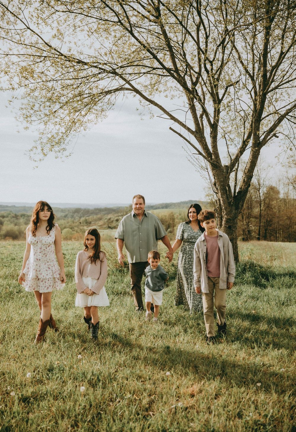 A family of six walks through a green meadow near a large tree on a cloudy spring day.indiana pa family photography, pittsburgh photographer