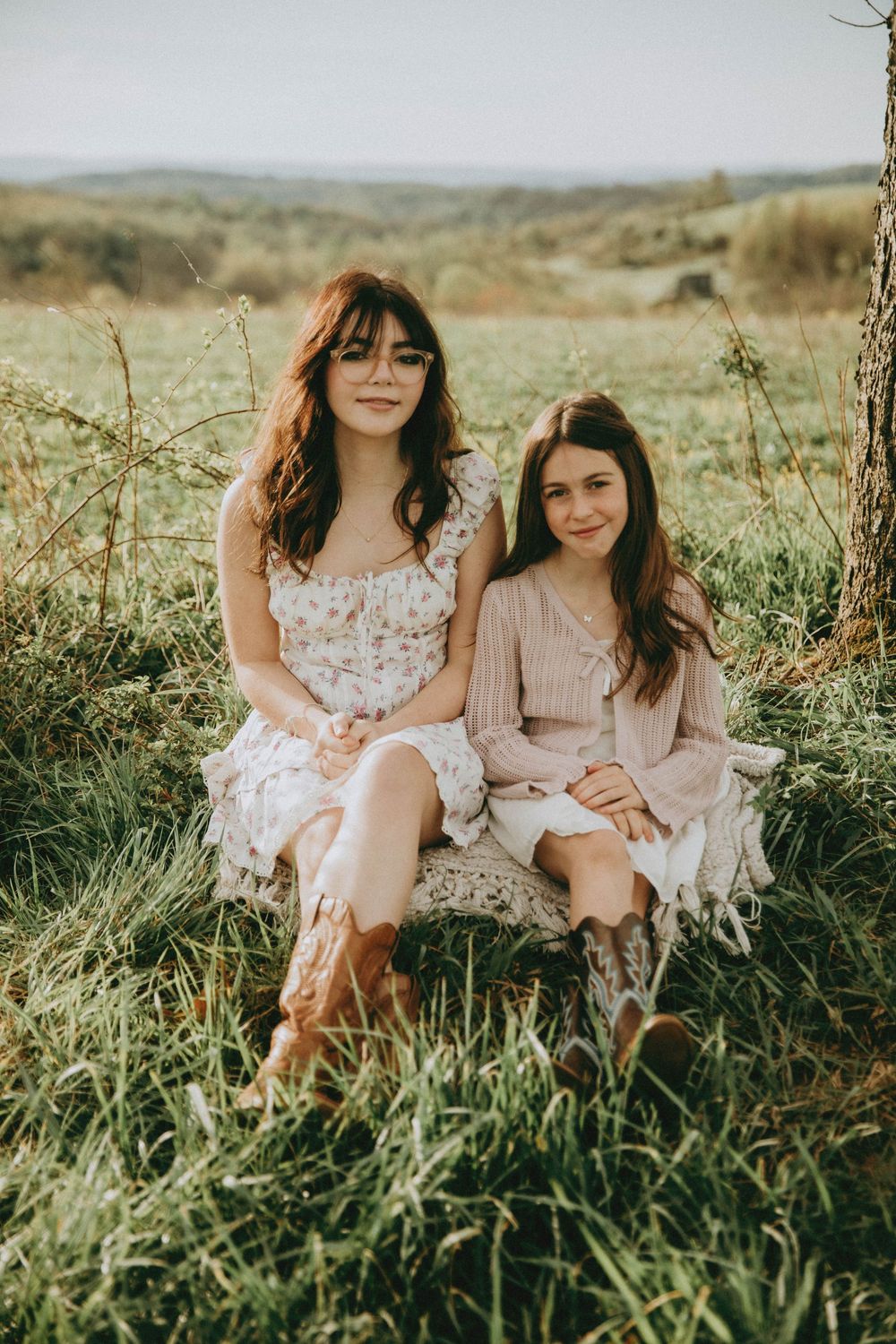 Two young women in floral dresses and cowboy boots sitting together in a green meadow on a sunny day. Indiana PA family photographer