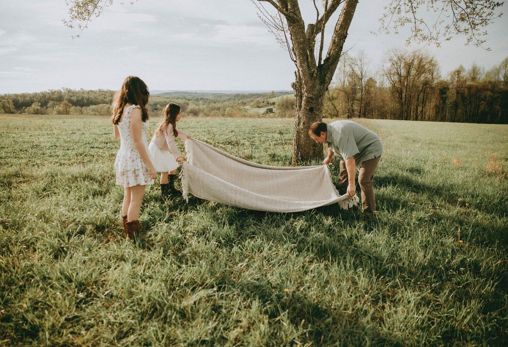 Three people spread a white blanket on lush green grass near a tree in a scenic countryside field. Indiana PA family photographer portraits