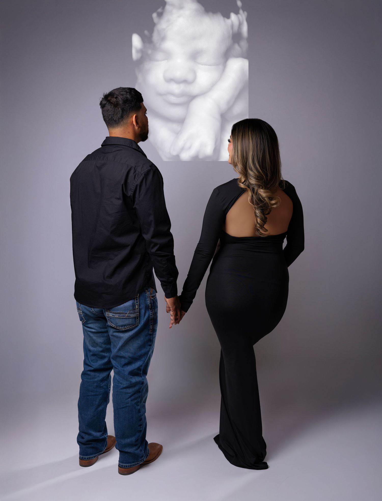 Couple in black holding hands facing a projected ultrasound image during a maternity photo shoot.