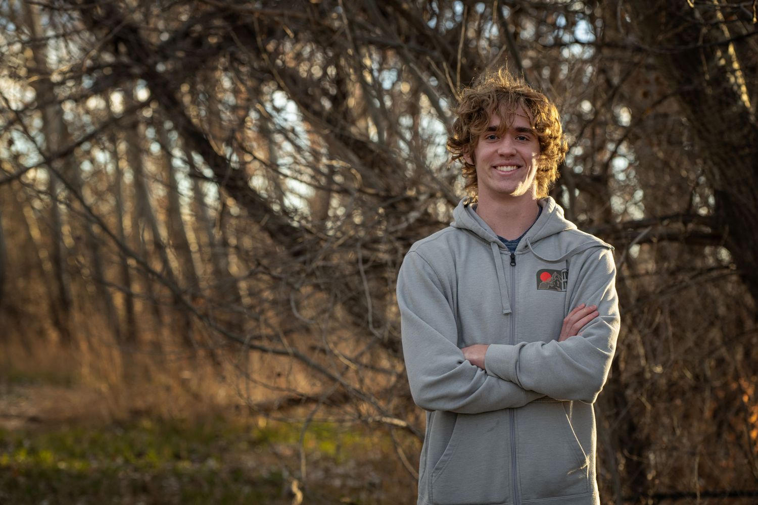 Young man with curly hair stands smiling outdoors with arms crossed, wearing a gray hoodie, surrounded by bare trees.