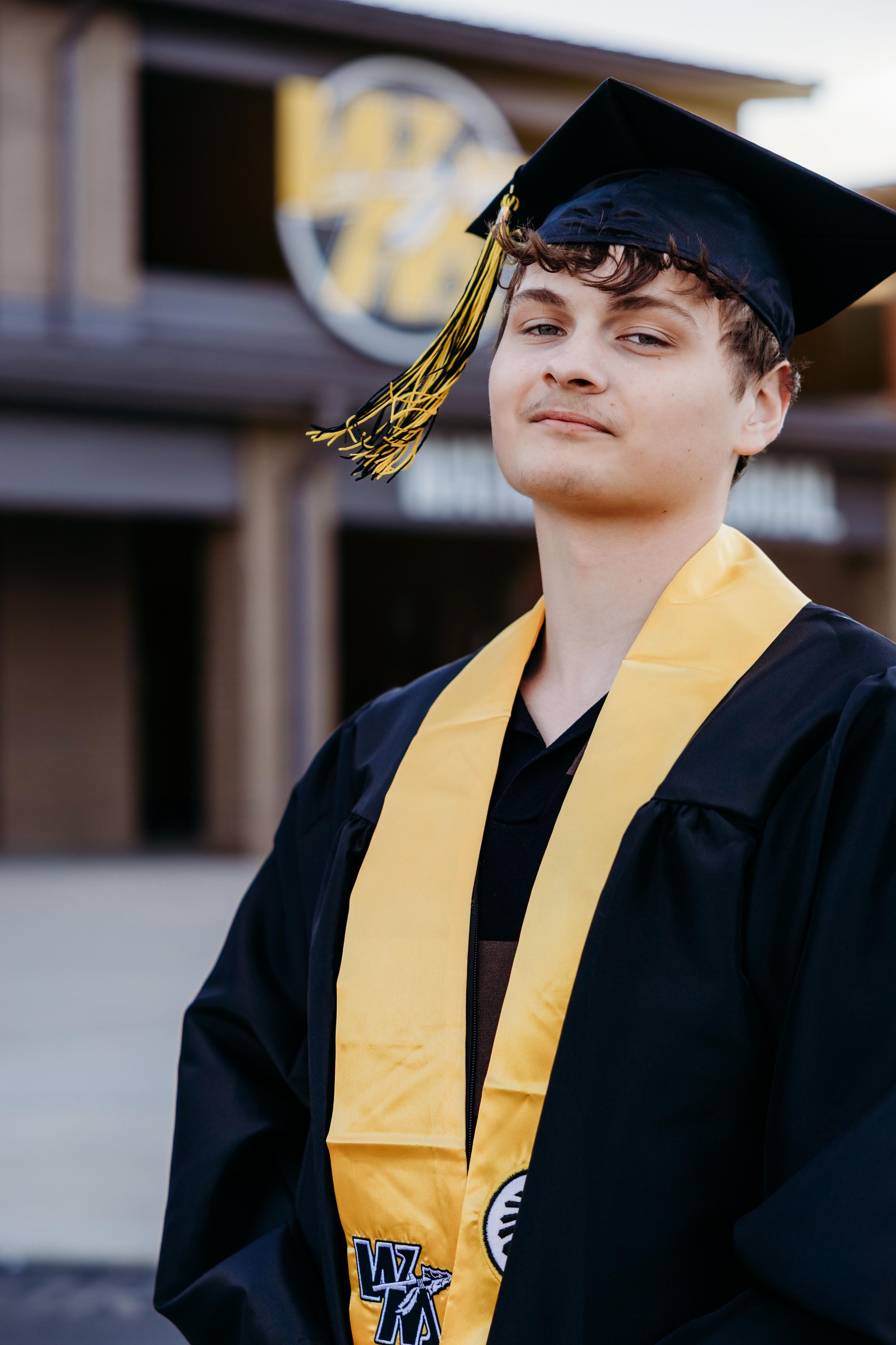 Young man in black graduation gown and cap with gold tassel and stole smiling outdoors at graduation ceremony.