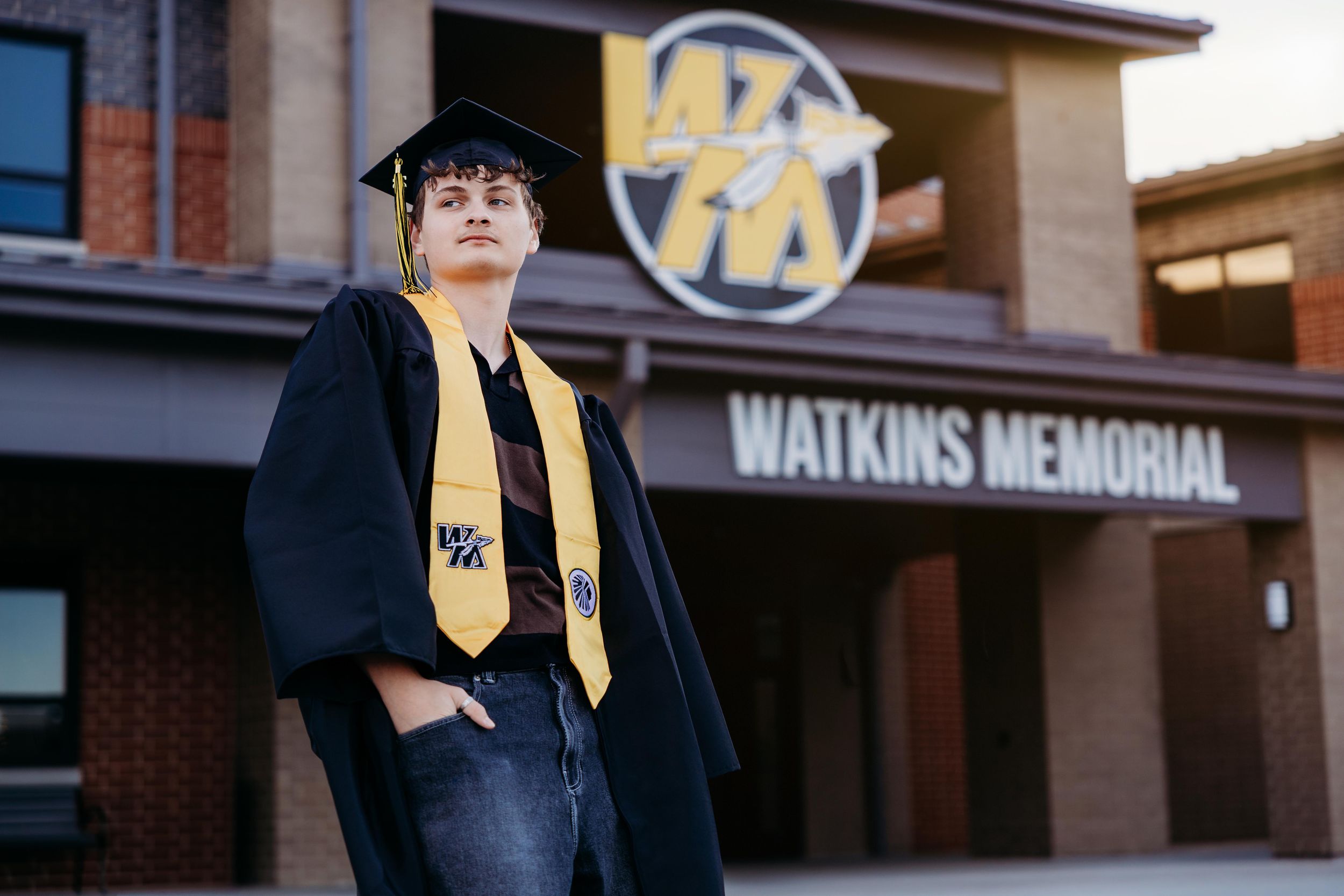 Graduate in cap and gown poses proudly in front of Watkins Memorial High School building.
