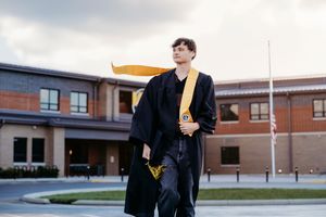 Young graduate in black cap and gown with gold stole stands proudly outside brick school building.
